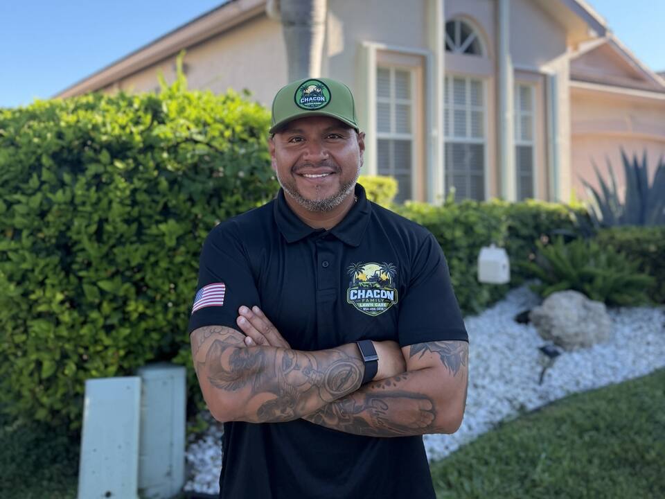 Juan Chacon, smiling with arms crossed, in his Chacon Family Lawn Care cap and polo in front of a South Florida home.
